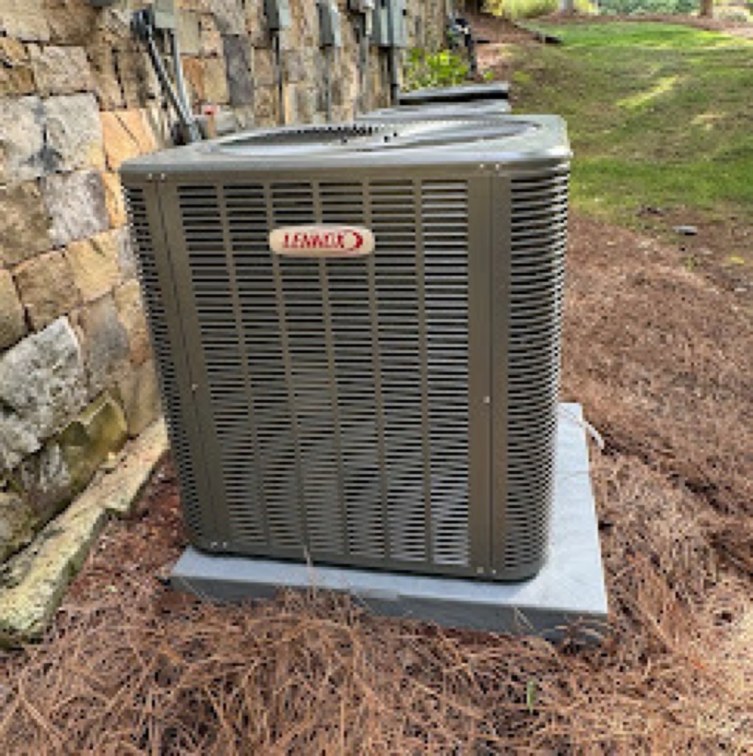 Lennox outdoor AC condenser unit installed on a concrete pad against a stone wall by Dynasty